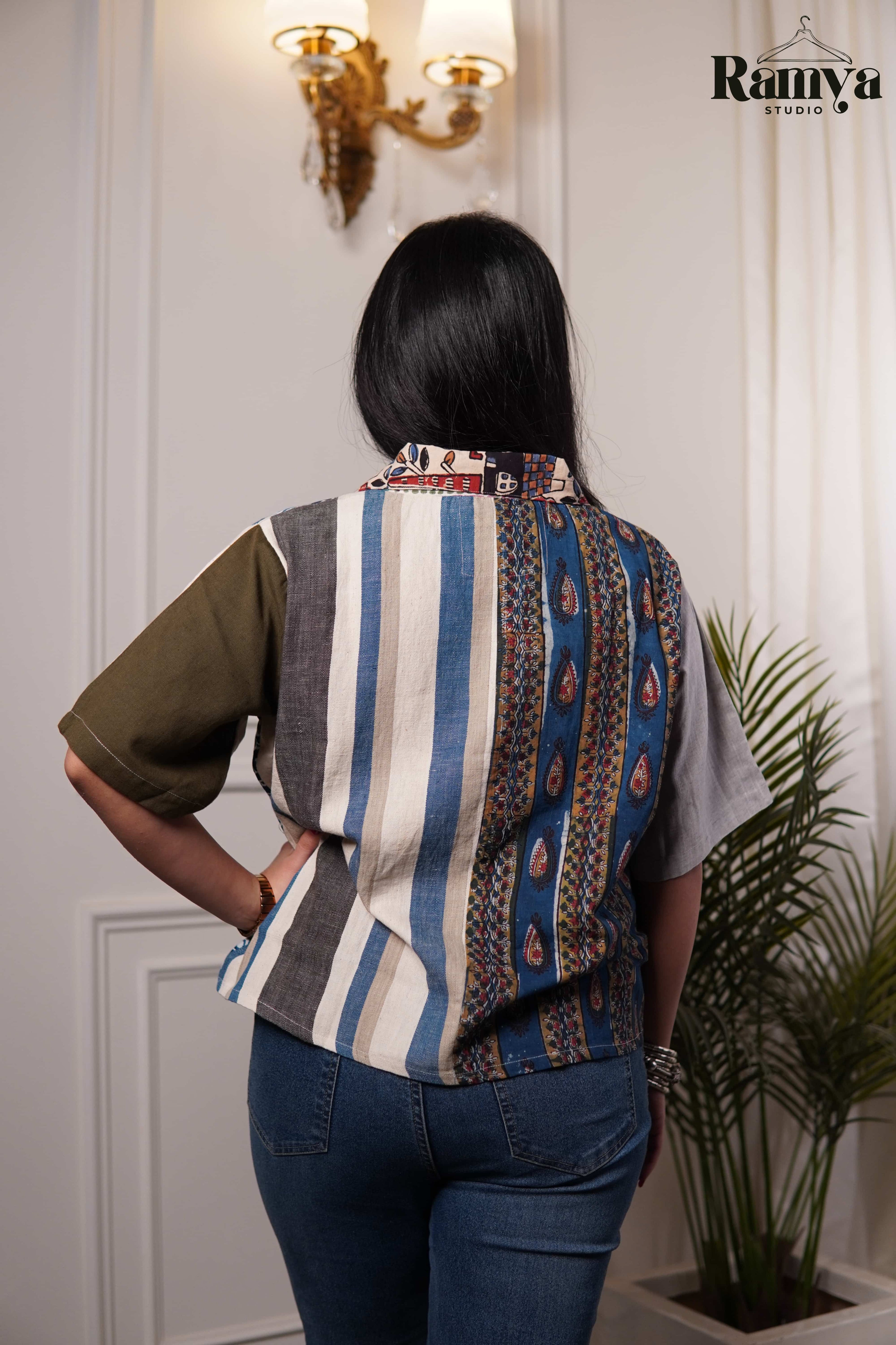 A woman wearing a short-sleeved button-down shirt with a beige, vibrant blue and white patterned panel, featuring a brown accent sleeve and accessorized with silver bangles.