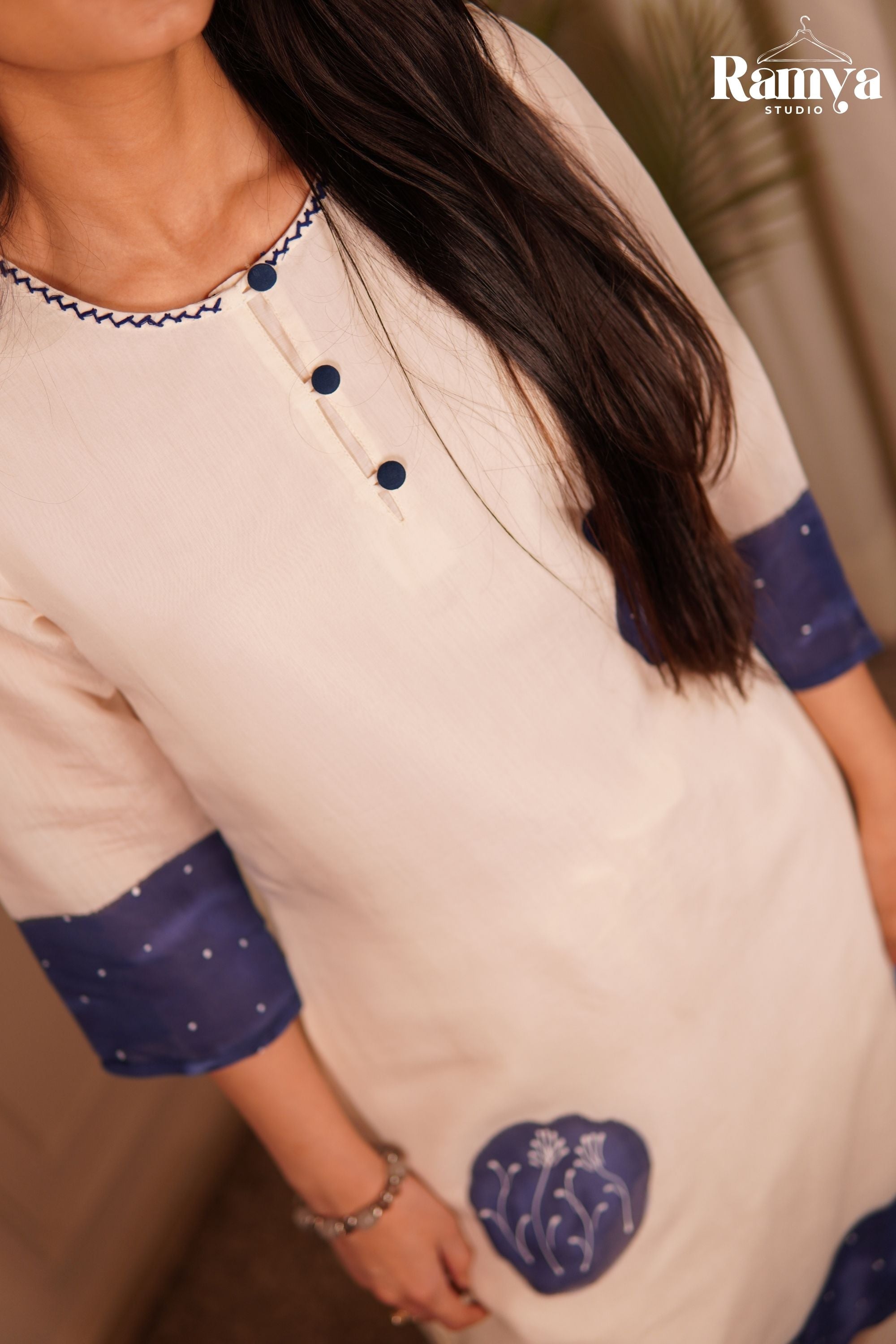 Close-up of the delicate white thread work embroidery inside an indigo applique patch on a cream tunic, highlighting matching fabric buttons and a fine dot print.