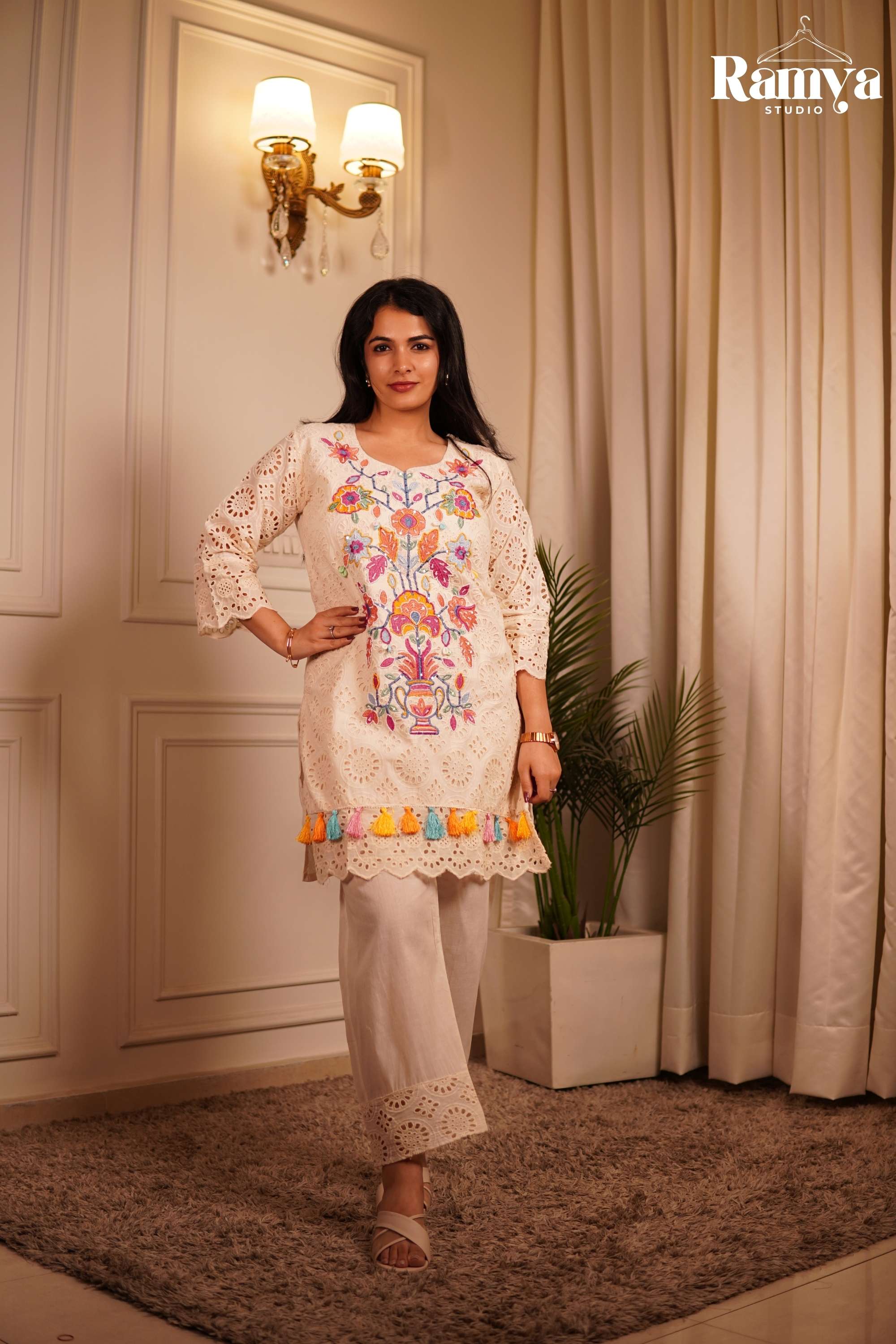 A model posing in Ramya Studio's 'Confetti on Cream' co-ord set, featuring an ivory Hakoba eyelet lace tunic with vibrant multicolored floral embroidery and pastel tassels.