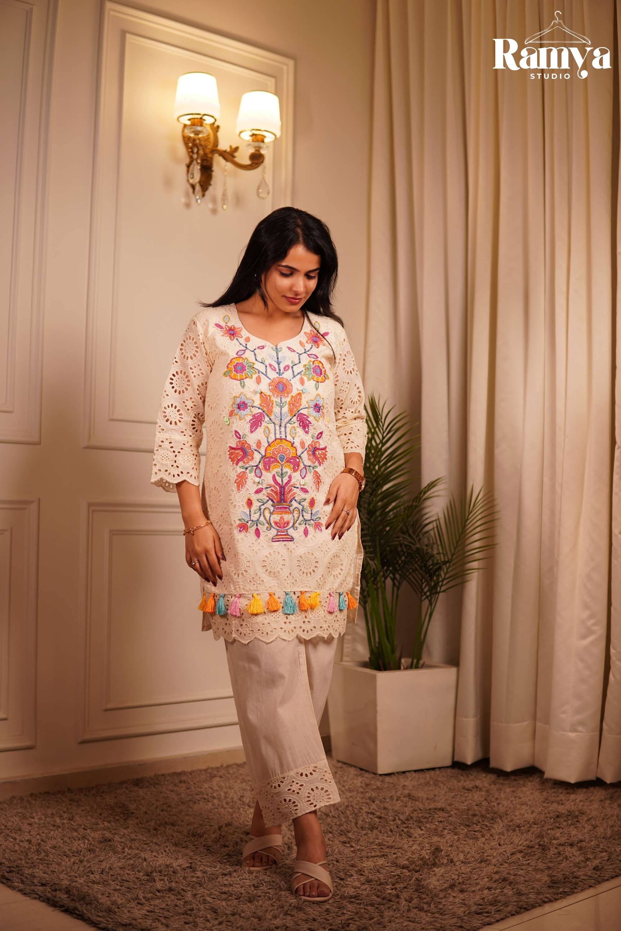 Boho-chic Indian resort wear: A model styling an ivory embroidered tunic set with cream cross-strap sandals against a luxurious minimalist home backdrop.