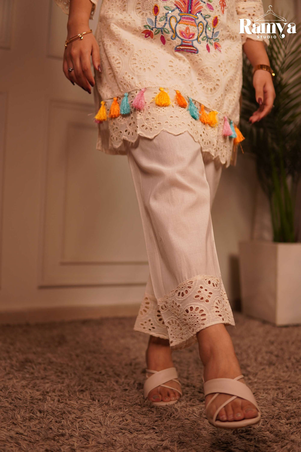 Close-up of a white eyelet lace short kurta with scalloped hems, colorful fringe thread tassels, and traditional botanical thread work on the yoke.