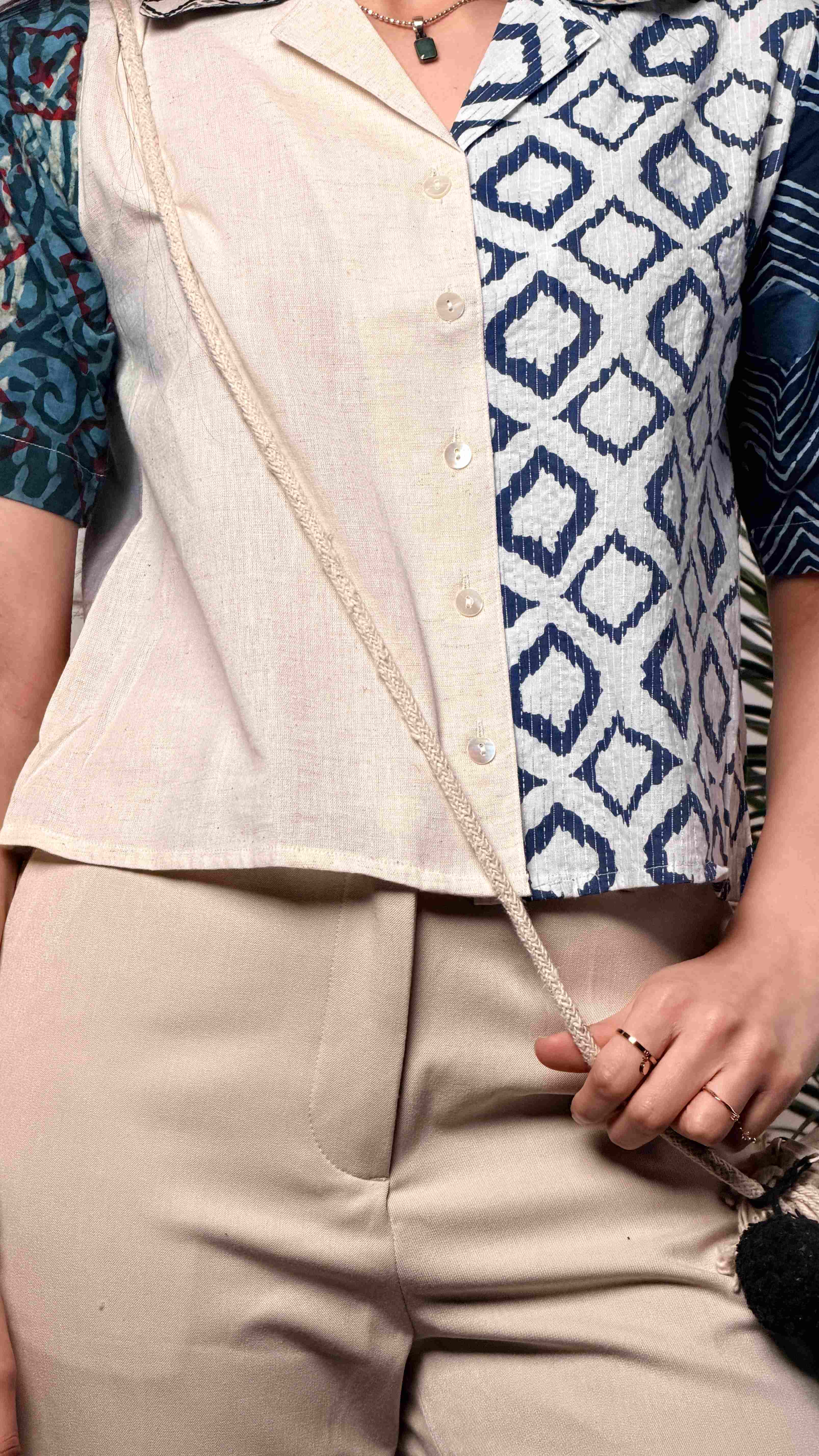 Woman wearing a button-up patchwork shirt with an off-white left panel and a blue diamond-patterned right panel. The shirt features a patterned collar and contrasting sleeves in teal and dark indigo prints.