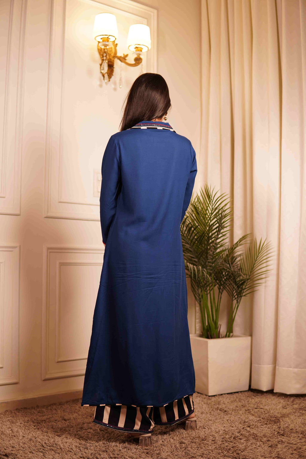 Woman wearing a blue co-ord set standing in a room with white walls and a plant.