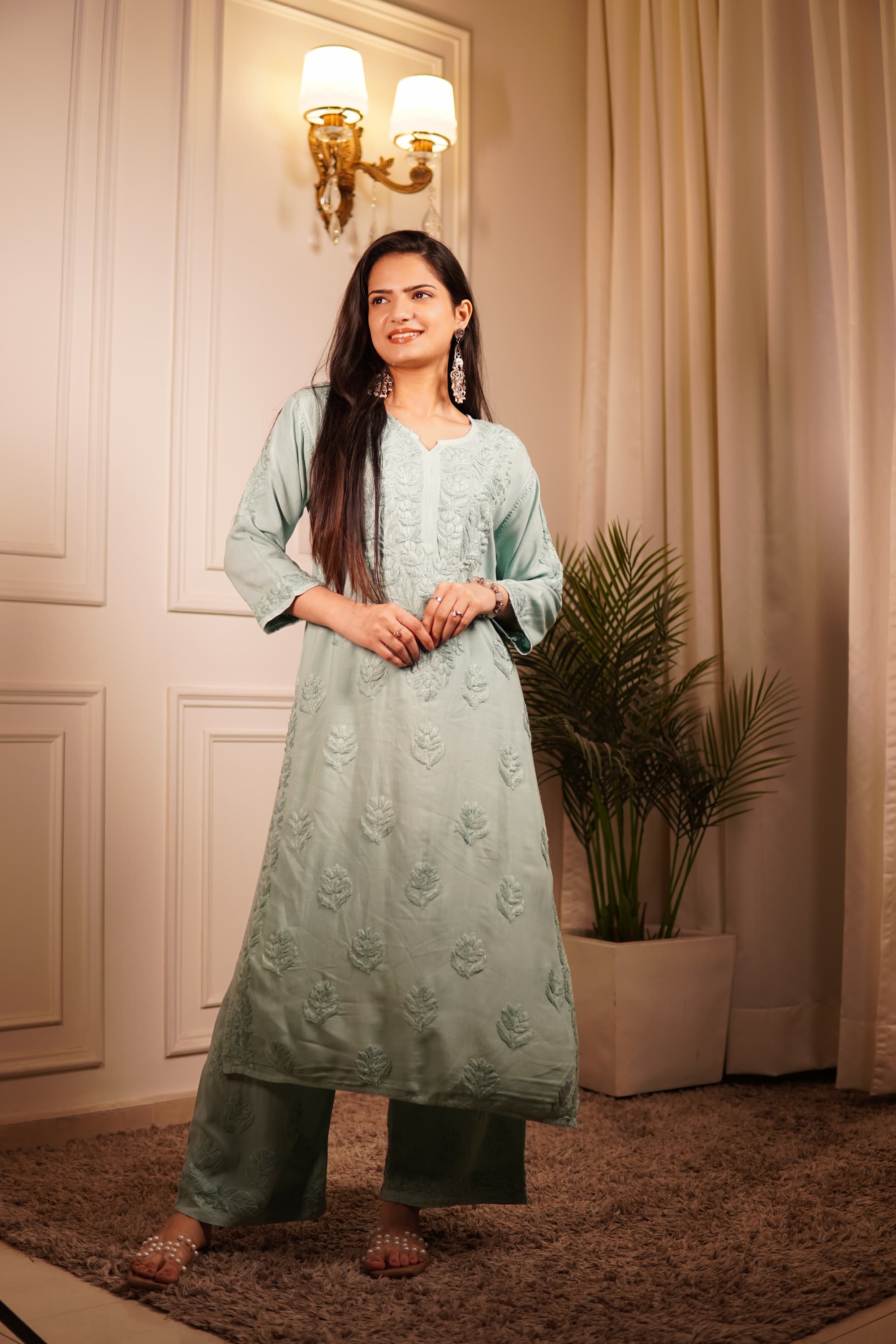 Woman in a light green kurta standing in a room with a plant and wall decor.