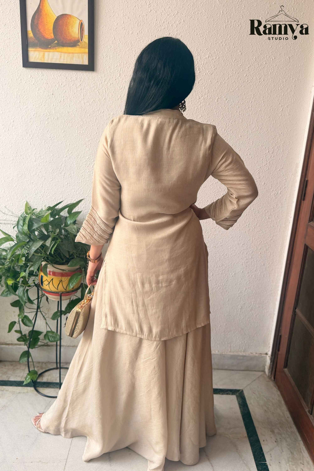 A woman is shown from behind wearing a beige long-sleeve kurta and matching flared skirt set, standing indoors against a white wall. She is holding a small beige clutch, with a potted plant on the left and a door on the right.