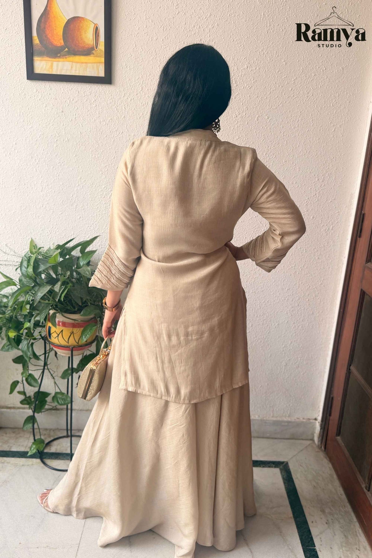 A woman is shown from behind wearing a beige long-sleeve kurta and matching flared skirt set, standing indoors against a white wall. She is holding a small beige clutch, with a potted plant on the left and a door on the right.