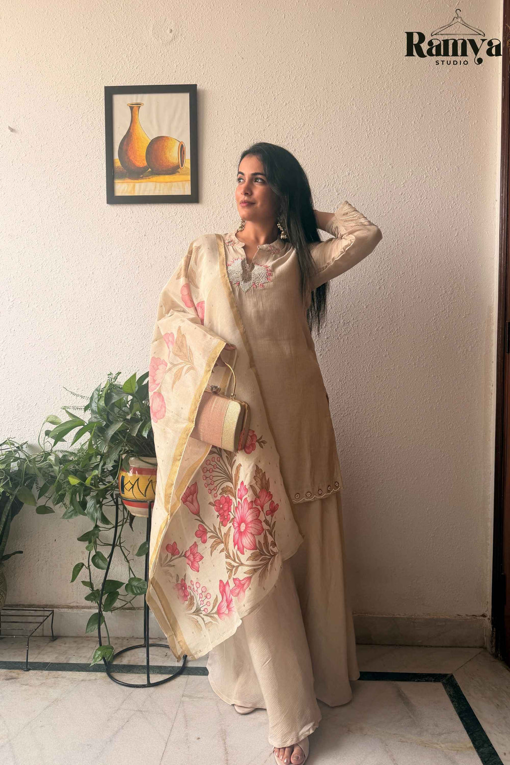 Full-length front view of a model wearing Ramya Studio's Geet Ivory Sharara Set holding a gold clutch and draped in a pink floral organza dupatta.