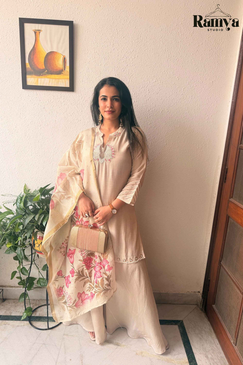 Full-length front view of a model wearing Ramya Studio's Geet Ivory Sharara Set holding a gold clutch and draped in a pink floral organza dupatta.