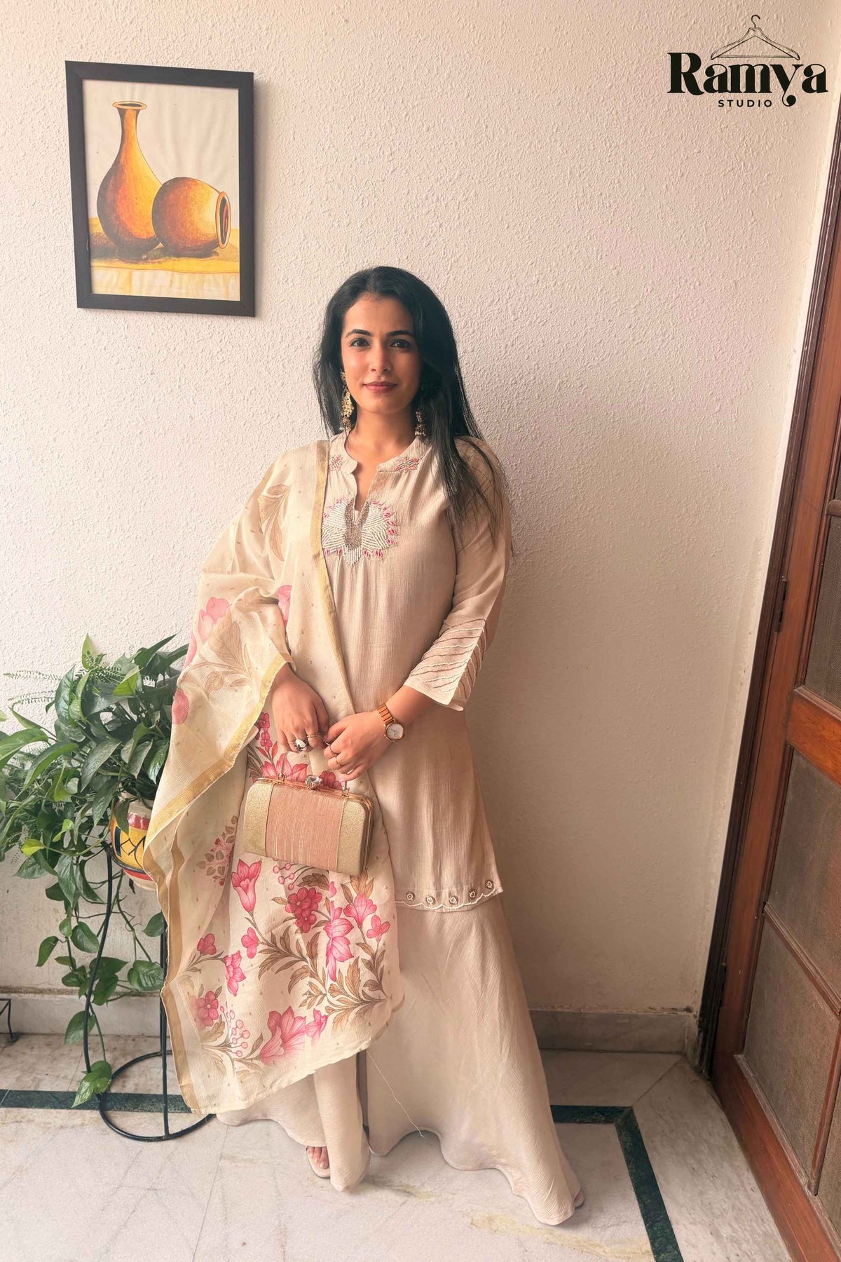 Full-length front view of a model wearing Ramya Studio's Geet Ivory Sharara Set holding a gold clutch and draped in a pink floral organza dupatta.