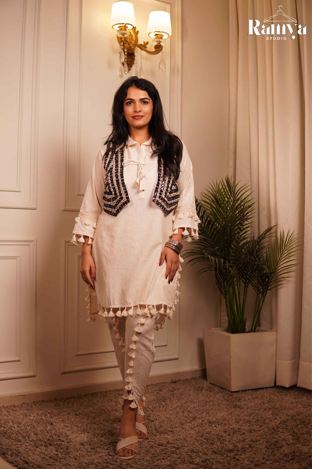 Full-length front view of a model wearing Ramya Studio's 'The Tassel-Tale Set', showcasing an off-white tunic with tassel fringes, a dark embroidered jacket, and tasseled tulip-slit pants.