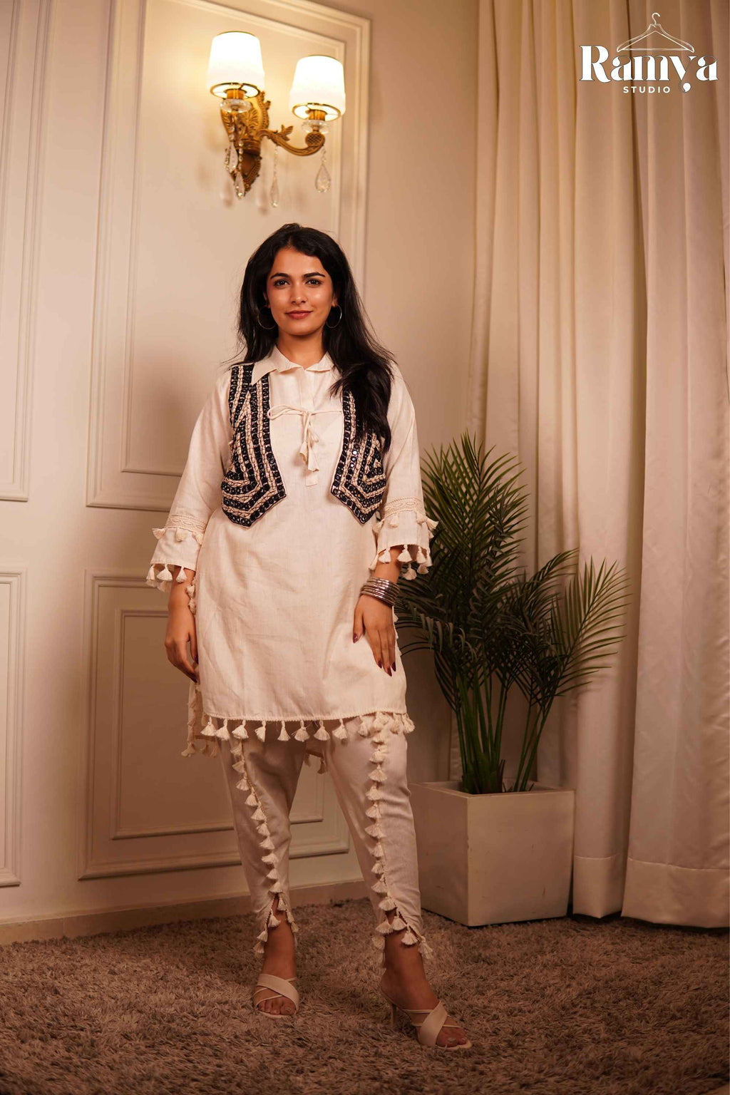 A model holding the tie-up drawstring on a navy geometric embroidered jacket paired with a tasseled ivory collared tunic and matching side-slit trousers.