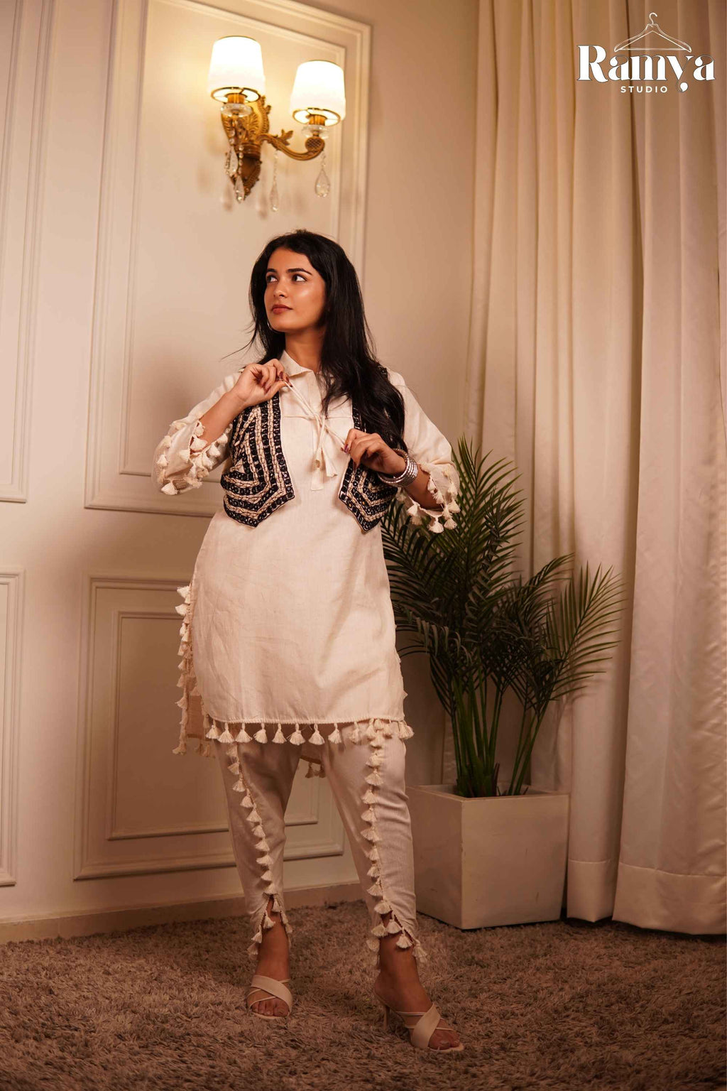 Full-length front view of a model in Ramya Studio's 'The Tassel-Tale Set', showing the ivory short tunic with fringe tassels, dark embroidered jacket koti, and matching tulip-slit trousers.