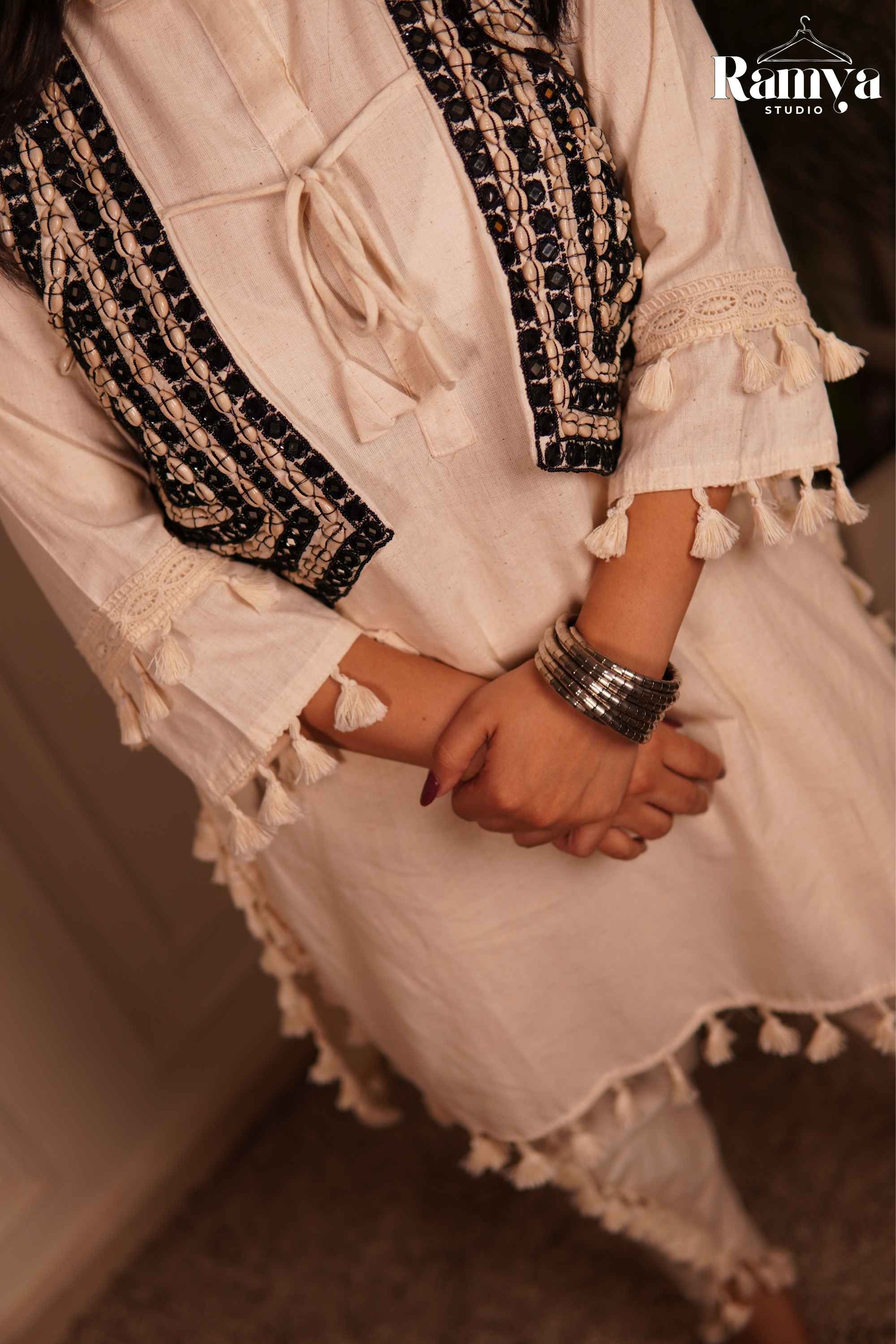 A close-up of a person wearing a cream-colored tunic with black and beige embroidered borders, lace trim, and tassels, accessorized with silver bangles. 